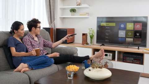兩個人並肩坐在客廳的沙發。一個人使用電視遙控器，瀏覽平面電視上顯示的直播服務選項選單