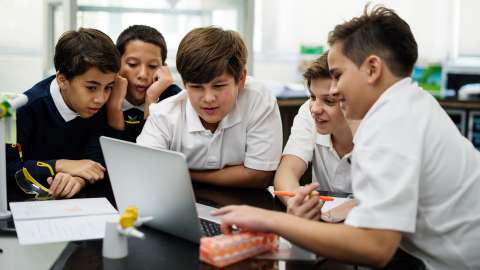 五名身穿校服的小學生，圍坐在教室環境中開機的筆記型電腦旁